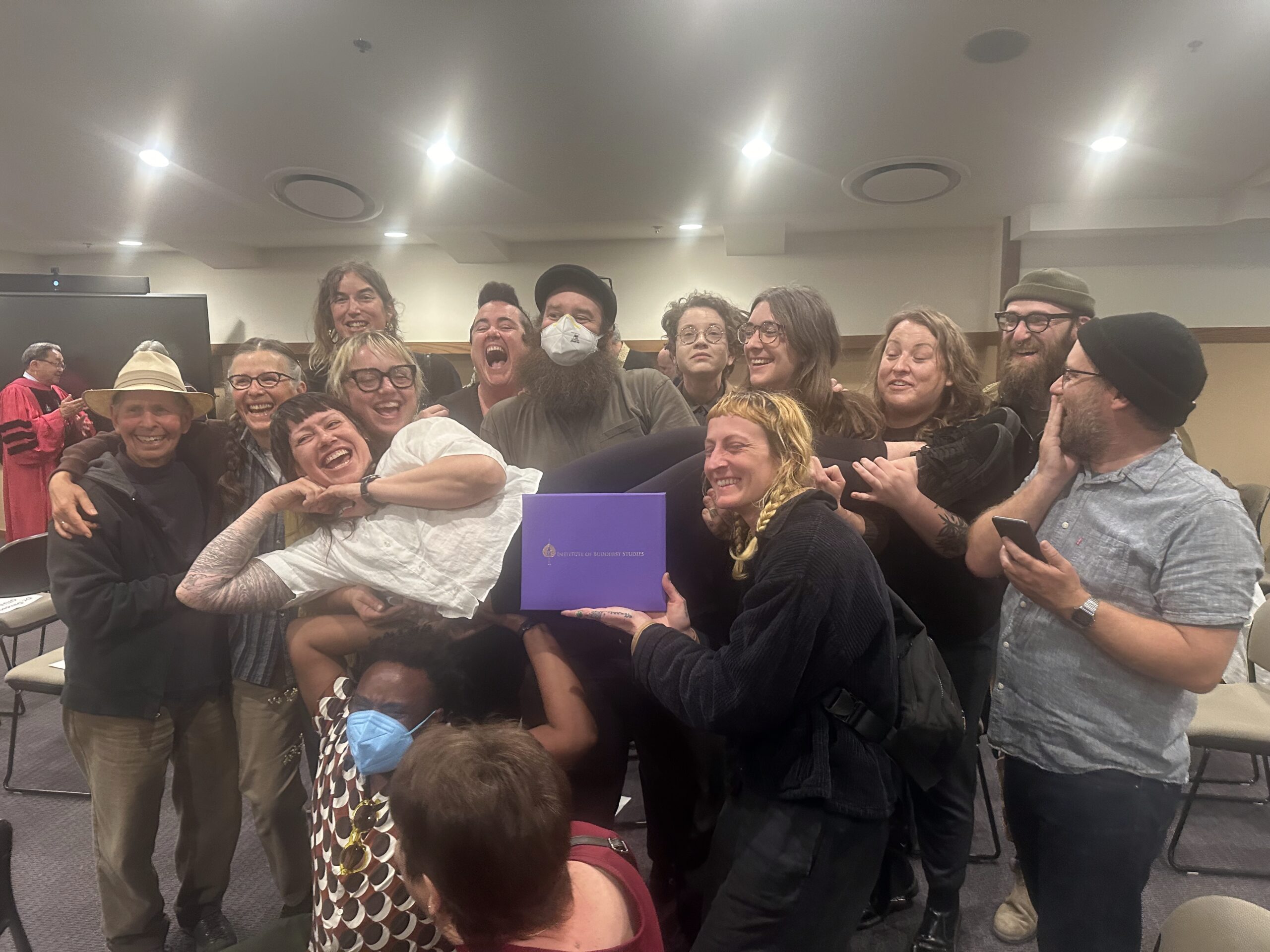Similing group of people, person in front holds purple envelope while remaining ground holds up a woman, laying on her side
